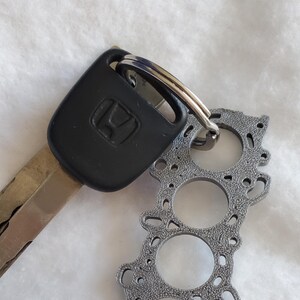 May include: A car key with a black Honda key fob attached to a silver metal keychain. The keychain has a grey, engine-themed pendant with three circular cutouts and detailed design.