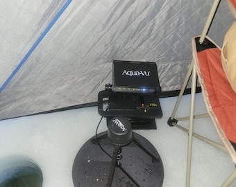 May include: Black Aqua-Vu underwater camera system with a round base and tripod legs, set up on ice. The screen displays the Aqua-Vu logo and model number 715c. A red and orange folding chair is in the background, and a gray tent is partially visible.