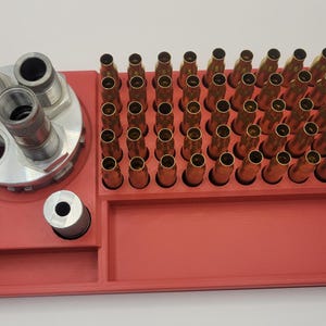 May include: A red reloading tray with multiple compartments. It holds brass casings and reloading tools. The tray has a rectangular shape with a section for tools and a section for holding the casings.