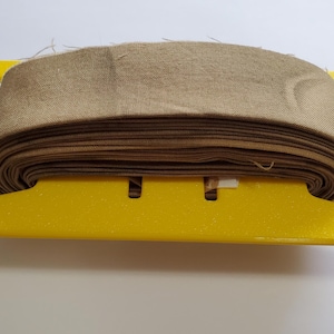 May include: A roll of beige fabric sits on a bright yellow spool holder. The fabric is tightly wound, showing multiple layers. The spool holder has a distinctive shape with extended arms.