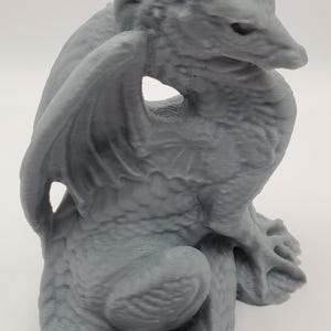 May include: A gray dragon figurine with detailed scales, wings, and a curled tail. The mythical creature is in a seated position, with its wings spread slightly. The dragon's head is turned to the side, and it has a textured surface.