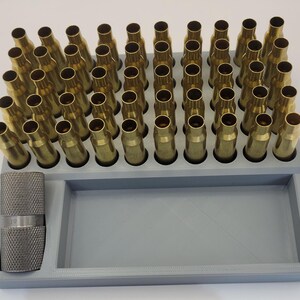 May include: A grey plastic reloading tray filled with brass cartridge casings. A textured metal tool is on the left. The tray is rectangular with holes for the casings and a larger open section.