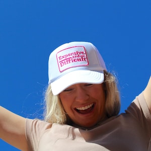 Op de afbeelding: Witte baseballcap met de tekst "Expensive and Difficult" in roze. De hoed heeft een vierkante patch op de voorkant. De achtergrond is een heldere blauwe lucht.