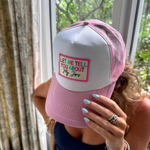 May include: Pink and white trucker hat with the text "LET ME TELL YOU ABOUT My Jesus" on a rectangular patch. The hat has a pink brim and mesh back. The text is in a colorful, multi-colored font.
