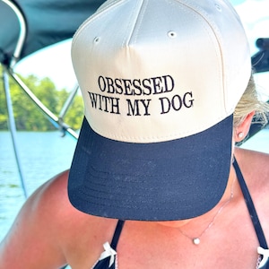 May include: A baseball cap with a cream-colored crown and a navy blue brim. The cap features the embroidered text "OBSESSED WITH MY DOG" in black. The cap has a black button on top.