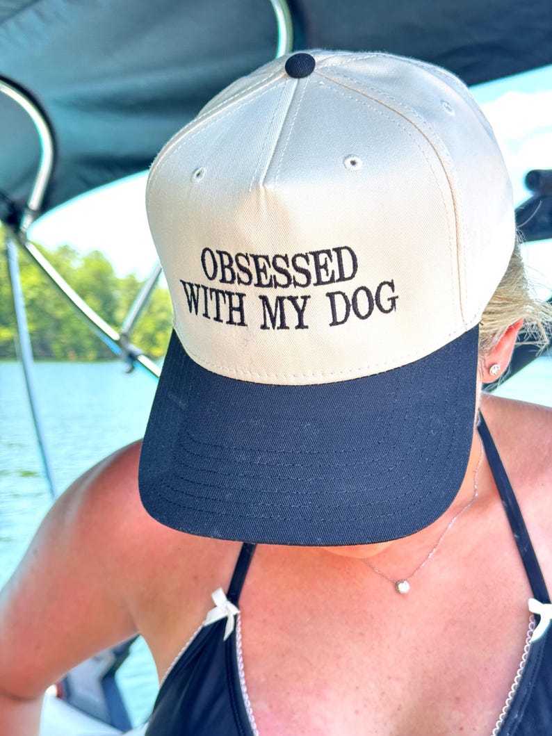 May include: A baseball cap with a cream-colored crown and a navy blue brim. The cap features the embroidered text "OBSESSED WITH MY DOG" in black. The cap has a black button on top.