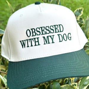 May include: A white and green baseball cap with the embroidered text "OBSESSED WITH MY DOG". The cap has a white crown and a dark green brim. The cap is a snapback style.