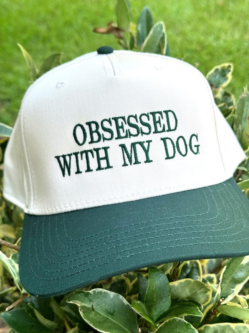 May include: A white and green baseball cap with the embroidered text "OBSESSED WITH MY DOG". The cap has a white crown and a dark green brim. The cap is a snapback style.