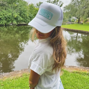 Puede incluir: Gorra de camionero blanca con parte trasera de malla y visera curva. La gorra tiene un parche rectangular con el texto "SOMEBODY'S PROBLEM" en color turquesa. La persona lleva una camisa blanca y vaqueros.