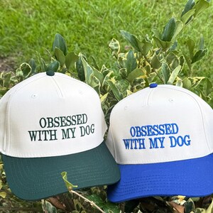May include: Two baseball caps with the text "OBSESSED WITH MY DOG". One cap has a white crown and a green brim, the other has a white crown and a blue brim. Both caps are embroidered with the text in matching colors.