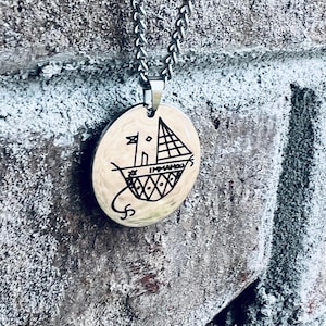 May include: A silver chain necklace with a round pendant featuring a black and white drawing of a sailboat. The pendant has a textured surface.