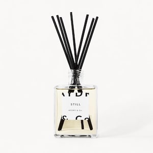 May include: A clear square glass reed diffuser bottle filled with light-colored liquid. It features a white label with black text reading STILL and AYDRY & Co. Several thin black reed sticks are inserted into the narrow neck of the bottle.
