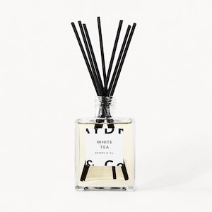 May include: A clear square glass reed diffuser bottle filled with light-colored liquid. The white label features the text "WHITE TEA" and "AYDRY & Co." in black font. Several thin black reeds are inserted into the bottle neck, fanning outward.