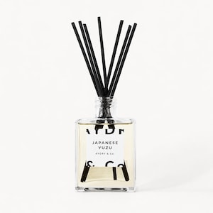 May include: A clear square glass reed diffuser bottle filled with light yellow liquid. The white label features black text reading AYDRY, JAPANESE YUZU, and AYDRY & Co. Seven thin black reeds are inserted into the bottle neck, fanning outward.