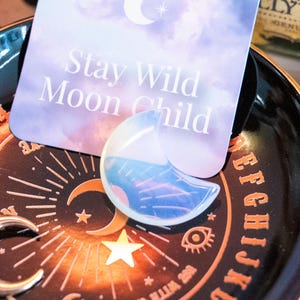 Könnte beinhalten: Ein schwarzes und goldenes Divinationsbrett mit einem Halbmond-Anhänger und einem regenbogenfarbenen, mondförmigen Kristall. Das Brett hat Buchstaben, Zahlen und Symbole. Die Karte im Hintergrund sagt "Stay Wild Moon Child".