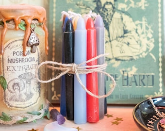 May include: A collection of candles and apothecary items. A bundle of colorful taper candles is tied with twine. A jar with a mushroom extract label and a mushroom charm is visible. A book and a decorative dish are in the background.