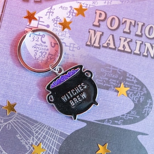 May include: A black cauldron keychain with purple glitter and the text "Witches Brew". The keychain is attached to a silver key ring.