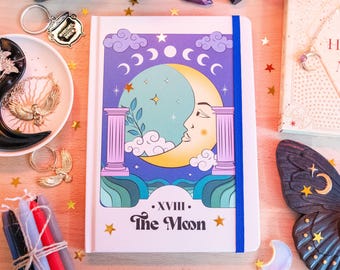 May include: A white journal with a blue elastic closure. The cover features a colorful illustration of a crescent moon with a face, surrounded by stars and clouds. The text "XVIII. The Moon" is printed in a retro font.