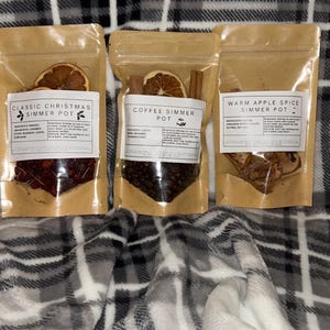 May include: Three clear pouches filled with simmer pot ingredients, each with a label. The pouches contain dried fruit, spices, and other botanicals. The labels read "Classic Christmas Simmer Pot", "Coffee Simmer Pot", and "Warm Apple Spice Simmer Pot".