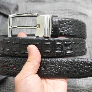 May include: A black leather belt with a crocodile-effect texture. The belt has a silver rectangular buckle. The belt is shown in the image.