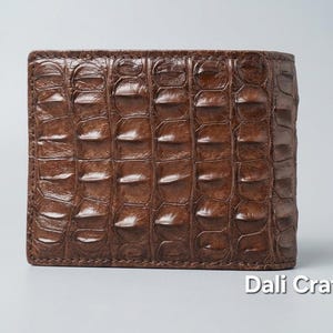 Puede incluir: Una cartera marrón de dos pliegues con un patrón de piel de cocodrilo texturizado y escamas en relieve. La superficie tiene un acabado brillante y un color marrón intenso. El texto Dali Craft Co está impreso en blanco en la esquina inferior.