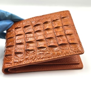 Può includere: Un portafoglio in pelle marrone con una texture a pelle di coccodrillo. Il portafoglio è rettangolare e ha un design a due ante. Il colore è un marrone rossastro caldo. Il portafoglio è tenuto da una mano guantata.