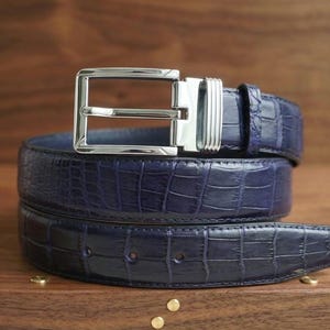 May include: A navy blue belt with a textured crocodile skin pattern and a polished silver-tone metal buckle. The belt is coiled in a circular shape, showcasing the embossed leather surface, the matching blue leather keeper, and the rectangular buckle.