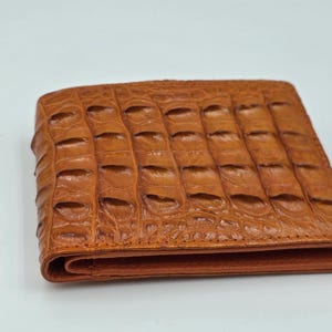 May include: A brown leather wallet with a crocodile skin texture. The rectangular wallet is folded, showing a seam and a subtle sheen. The colour is a warm, medium brown.