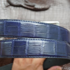 May include: A close-up of a blue leather belt with a crocodile pattern. The belt has a silver buckle and is made of a textured, high-quality material. The belt is approximately 3.8 cm wide.
