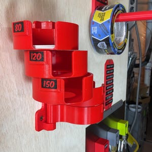 May include: A red wall-mounted organizer featuring four stacked circular holders labeled 80, 120, and 150 in black text. A red horizontal rod extends from a separate red mount holding a roll of blue and yellow painter's tape with ProSharp text.