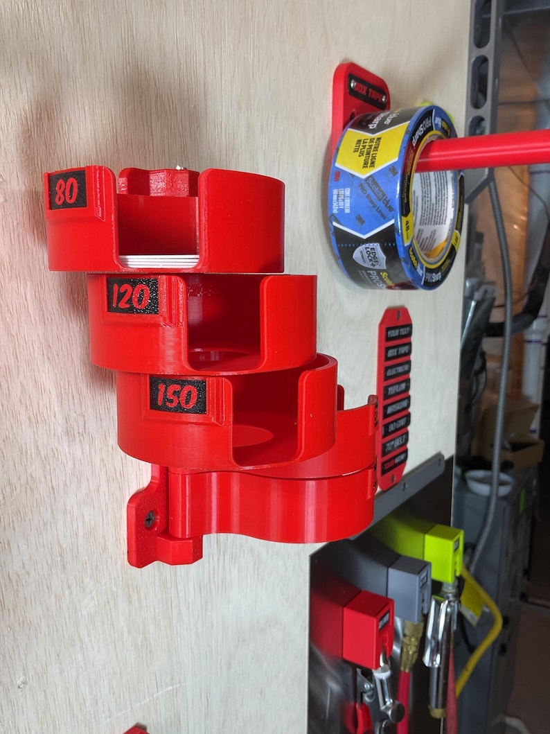 May include: A red wall-mounted organizer featuring four stacked circular holders labeled 80, 120, and 150 in black text. A red horizontal rod extends from a separate red mount holding a roll of blue and yellow painter's tape with ProSharp text.