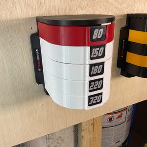 Puede incluir: Un estante de almacenamiento montado en la pared con una base negra y cinco compartimentos curvos apilados. El compartimento superior es rojo y los cuatro inferiores son blancos. Cada uno tiene una etiqueta negra con n&uacute;meros blancos: 80, 150, 180, 220 y 320. El lateral tiene la marca TOUGH-GEAR.