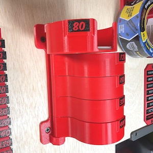 May include: A red wall-mounted organizer for sanding discs featuring five stacked circular holders. Each holder is labeled with black text indicating grit levels 80, 120, 150, 320, and 400. The unit is secured to a light wood surface with two visible screws.