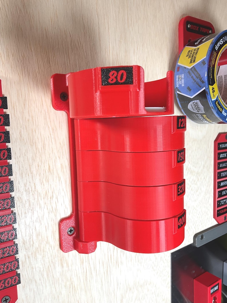 May include: A red wall-mounted organizer for sanding discs featuring five stacked circular holders. Each holder is labeled with black text indicating grit levels 80, 120, 150, 320, and 400. The unit is secured to a light wood surface with two visible screws.