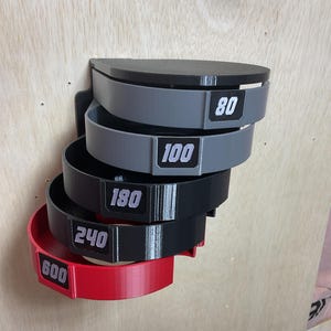 Puede incluir: Un organizador de pared con cinco soportes semicirculares apilados en orden descendente. Los soportes son de color gris, negro y rojo, cada uno etiquetado con los n&uacute;meros blancos 80, 100, 180, 240 y 600 sobre un fondo rectangular negro.
