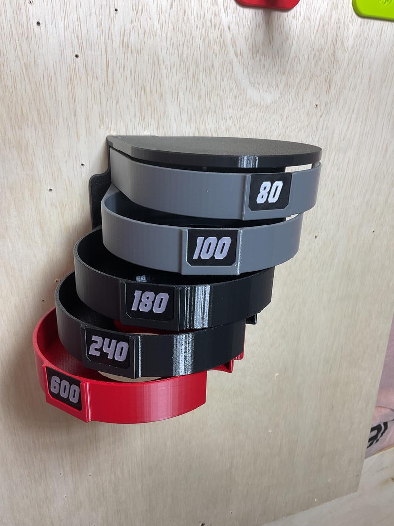 Puede incluir: Un organizador de pared con cinco soportes semicirculares apilados en orden descendente. Los soportes son de color gris, negro y rojo, cada uno etiquetado con los n&uacute;meros blancos 80, 100, 180, 240 y 600 sobre un fondo rectangular negro.