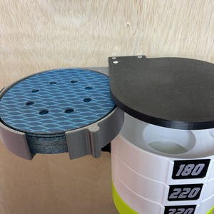 Puede incluir: Un organizador de pared que incluye un soporte gris para discos de lija azules con patr&oacute;n de diamante y una torre cil&iacute;ndrica blanca con tapa negra para almacenar granos, etiquetada con los n&uacute;meros 180, 220, 320 y 400 en color negro.
