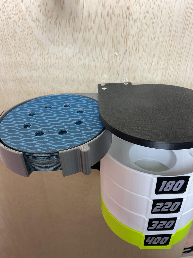 Puede incluir: Un organizador de pared que incluye un soporte gris para discos de lija azules con patr&oacute;n de diamante y una torre cil&iacute;ndrica blanca con tapa negra para almacenar granos, etiquetada con los n&uacute;meros 180, 220, 320 y 400 en color negro.