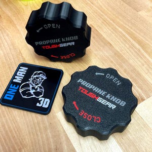 May include: Two black circular propane knobs with scalloped edges, featuring the text "PROPANE KNOB TOUGHGEAR" in white and red, with "OPEN" and "CLOSE" arrows. A square black sticker with a blue "ONE MAN 3D" logo and a cartoon character.