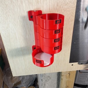 May include: A bright red, wall-mounted storage rack designed for circular sanding discs. The unit features five stacked cylindrical compartments, each labeled with black text indicating grit sizes 80, 120, 150, 320, and 400.