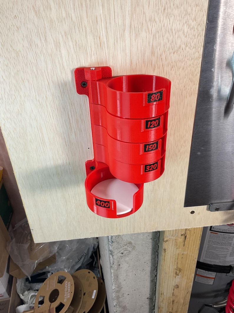 May include: A bright red, wall-mounted storage rack designed for circular sanding discs. The unit features five stacked cylindrical compartments, each labeled with black text indicating grit sizes 80, 120, 150, 320, and 400.