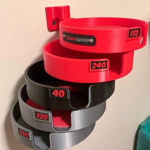 May include: A wall-mounted storage rack featuring four stacked circular holders in red, black, and gray. Each holder displays a grit number label: 150, 240, 40, 220, and 320. The top red holder features a black plate with the text TOUGHGEAR.