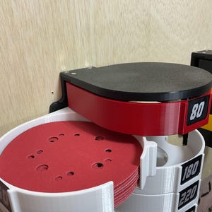 Puede incluir: Un organizador de pared para discos de lijado circulares con una secci&oacute;n superior roja etiquetada 80 y secciones inferiores blancas etiquetadas 180, 220 y 320. La unidad sostiene una pila de discos de lijado rojos con orificios visibles.