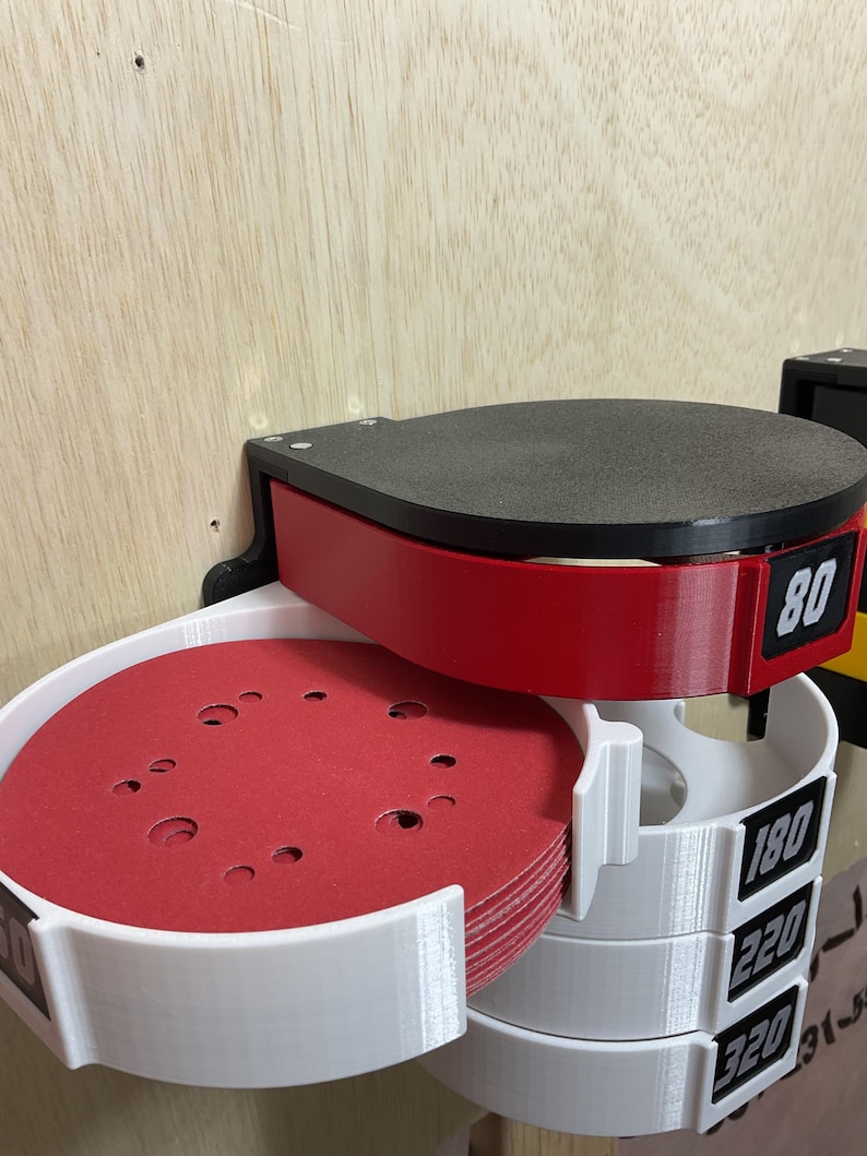 Puede incluir: Un organizador de pared para discos de lijado circulares con una secci&oacute;n superior roja etiquetada 80 y secciones inferiores blancas etiquetadas 180, 220 y 320. La unidad sostiene una pila de discos de lijado rojos con orificios visibles.