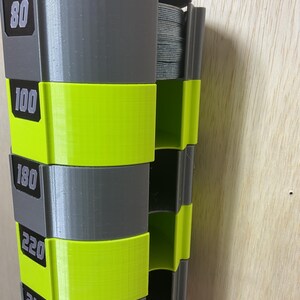 Puede incluir: Un dispensador de almacenamiento vertical montado en la pared con segmentos alternos en gris y amarillo ne&oacute;n. Las secciones grises est&aacute;n etiquetadas con los n&uacute;meros 80, 100, 180, 220 y 240. La unidad contiene una pila de discos de lija grises.