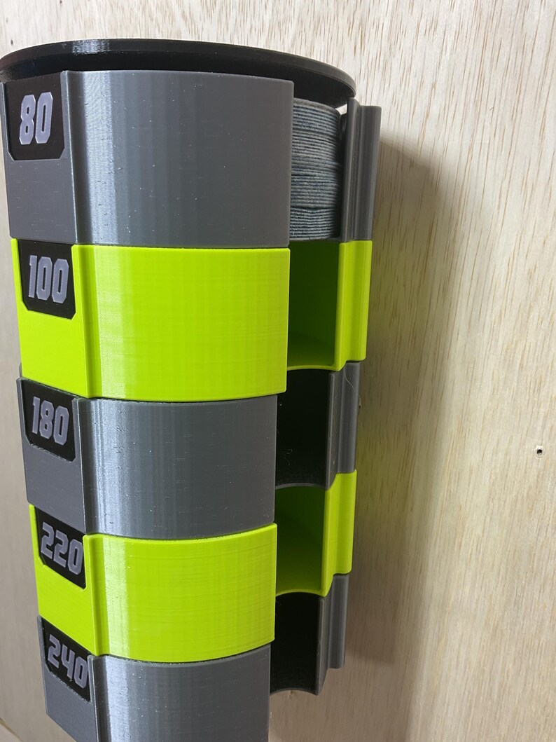 Puede incluir: Un dispensador de almacenamiento vertical montado en la pared con segmentos alternos en gris y amarillo ne&oacute;n. Las secciones grises est&aacute;n etiquetadas con los n&uacute;meros 80, 100, 180, 220 y 240. La unidad contiene una pila de discos de lija grises.
