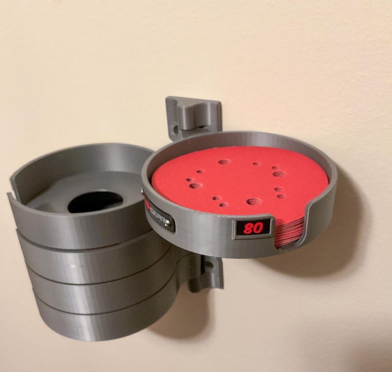 May include: A gray wall-mounted storage organizer for circular sanding discs. The unit features a stack of gray cylindrical holders with a prominent red sanding disc visible in the top compartment, which is clearly labeled with the number 80 in red.