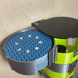 Puede incluir: Un organizador de pared para discos de lija circulares, con un marco de pl&aacute;stico gris y una pila de discos abrasivos de malla azul. La unidad incluye una tapa negra y una secci&oacute;n vertical de color amarillo brillante con las etiquetas 100, 180, 220 y 240.