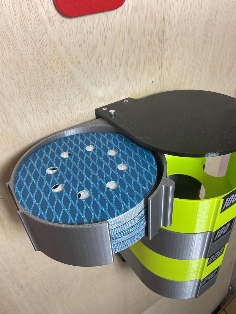 Puede incluir: Un organizador de pared para discos de lija circulares, con un marco de pl&aacute;stico gris y una pila de discos abrasivos de malla azul. La unidad incluye una tapa negra y una secci&oacute;n vertical de color amarillo brillante con las etiquetas 100, 180, 220 y 240.