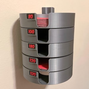 May include: A gray wall-mounted storage organizer for circular sanding discs, featuring five stacked compartments. Each section has a U-shaped cutout and a black label with red text indicating grit levels 80, 100, 180, 220, and 320.
